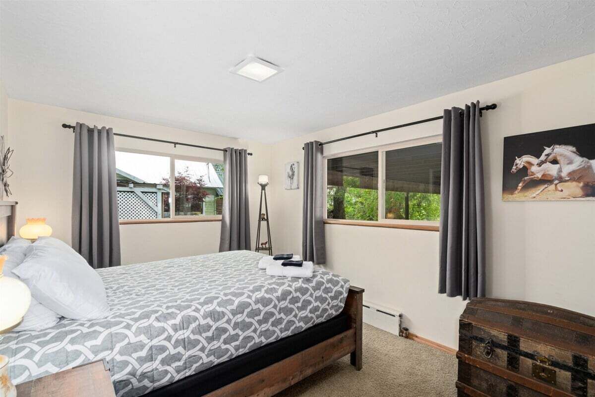 Une chambre à coucher avec un lit, deux fenêtres avec des rideaux, une lampe, une échelle, un tableau et une commode.