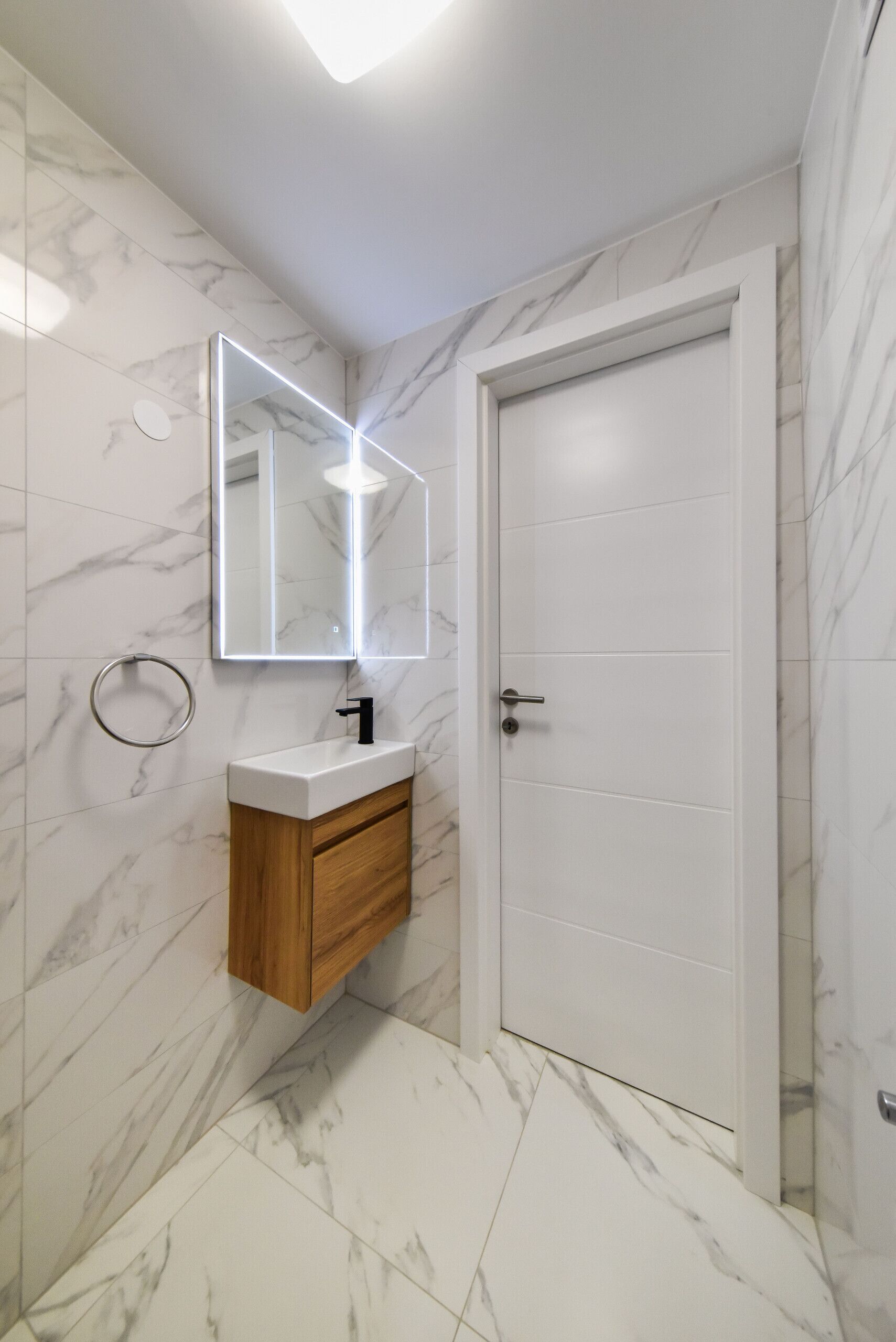 Une salle de bain avec un sol carrelé de marbre, un meuble-lavabo en bois avec un robinet noir, un miroir rond avec un éclairage intégré et une porte blanche.