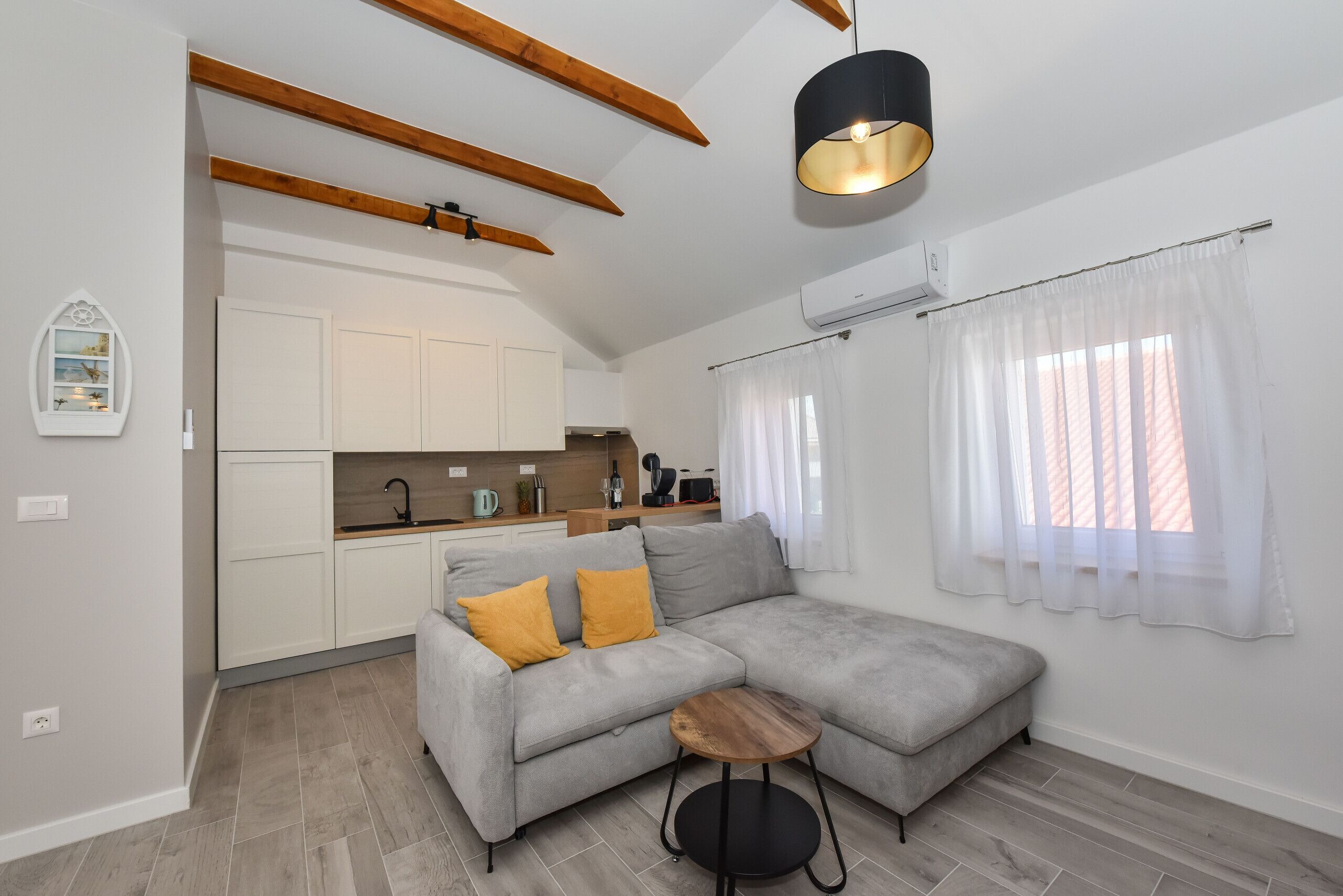 Un salón moderno con un sofá seccional gris, una mesita de madera y una lámpara colgante. Se observa un área de cocina con armarios blancos y una encimera de madera al fondo.
