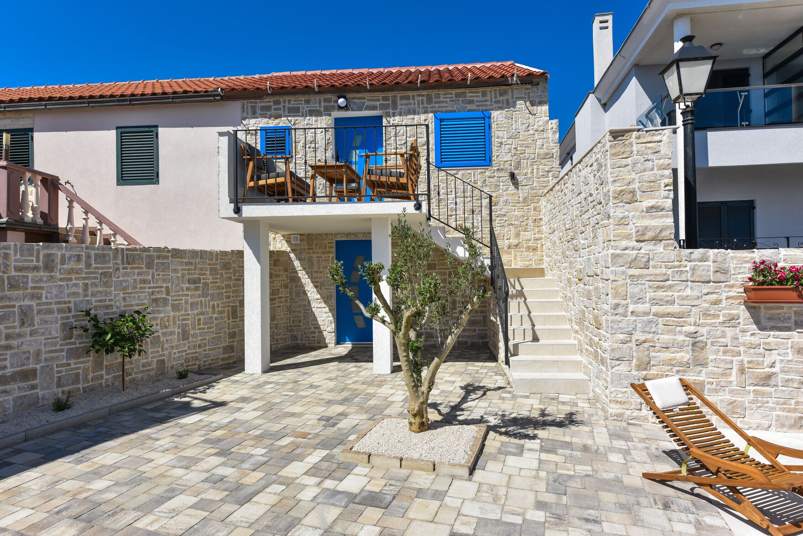 A courtyard with a stone wall, a blue door, and a staircase leading to a balcony with wooden furniture.