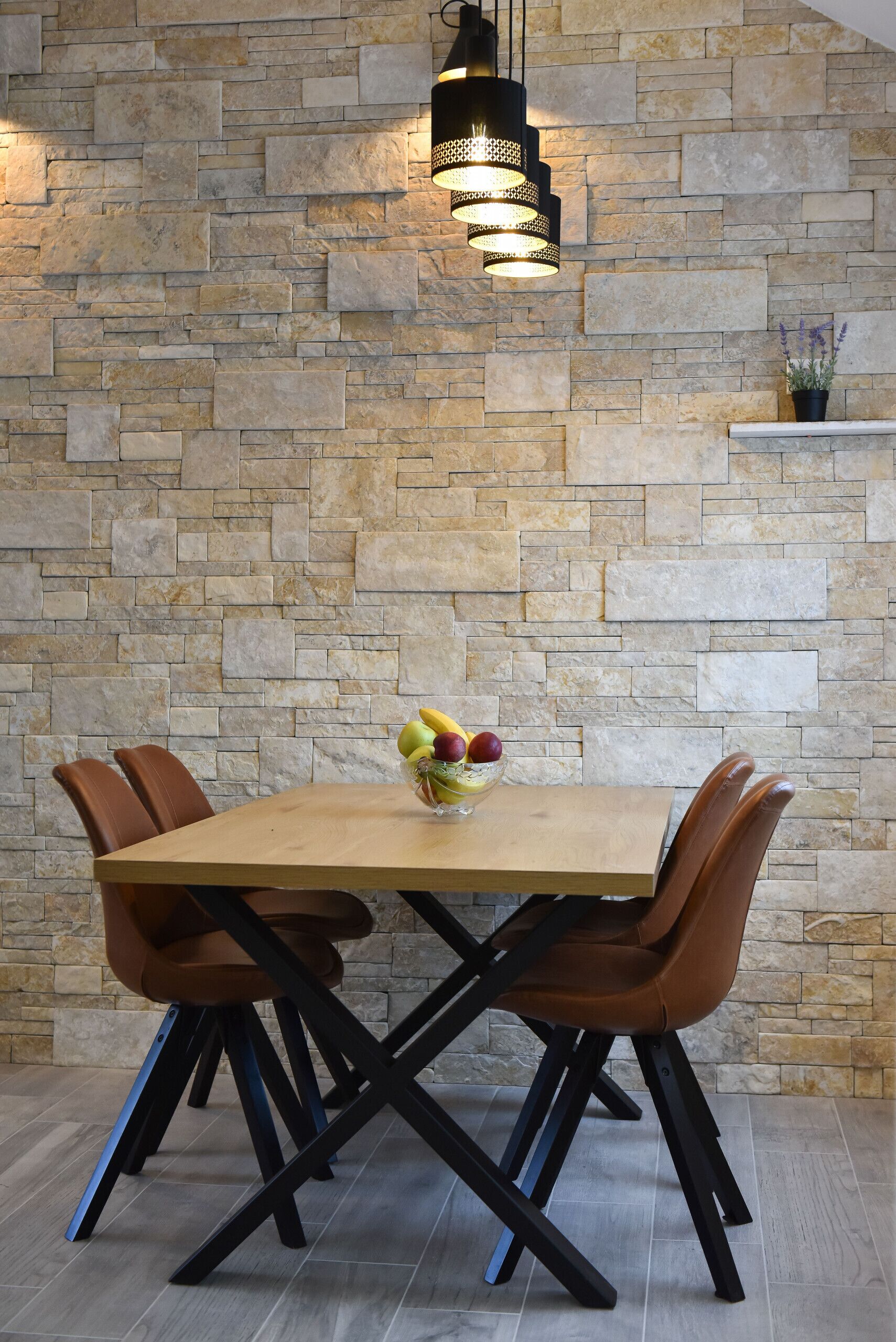 A dining table with a fruit bowl, surrounded by chairs, against a stone wall with a hanging light fixture.