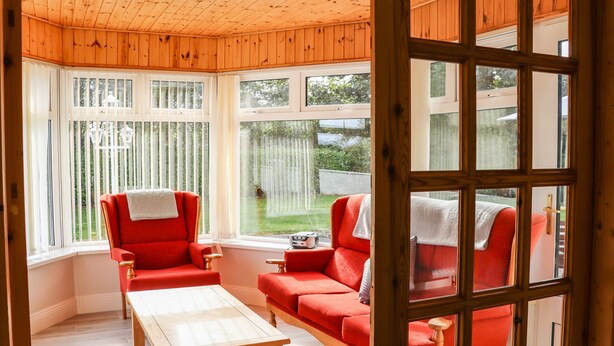 A room with wooden paneled walls and ceiling, a red sofa set, a wooden table, and large windows with white blinds. - Woodlawn (Kilmore)