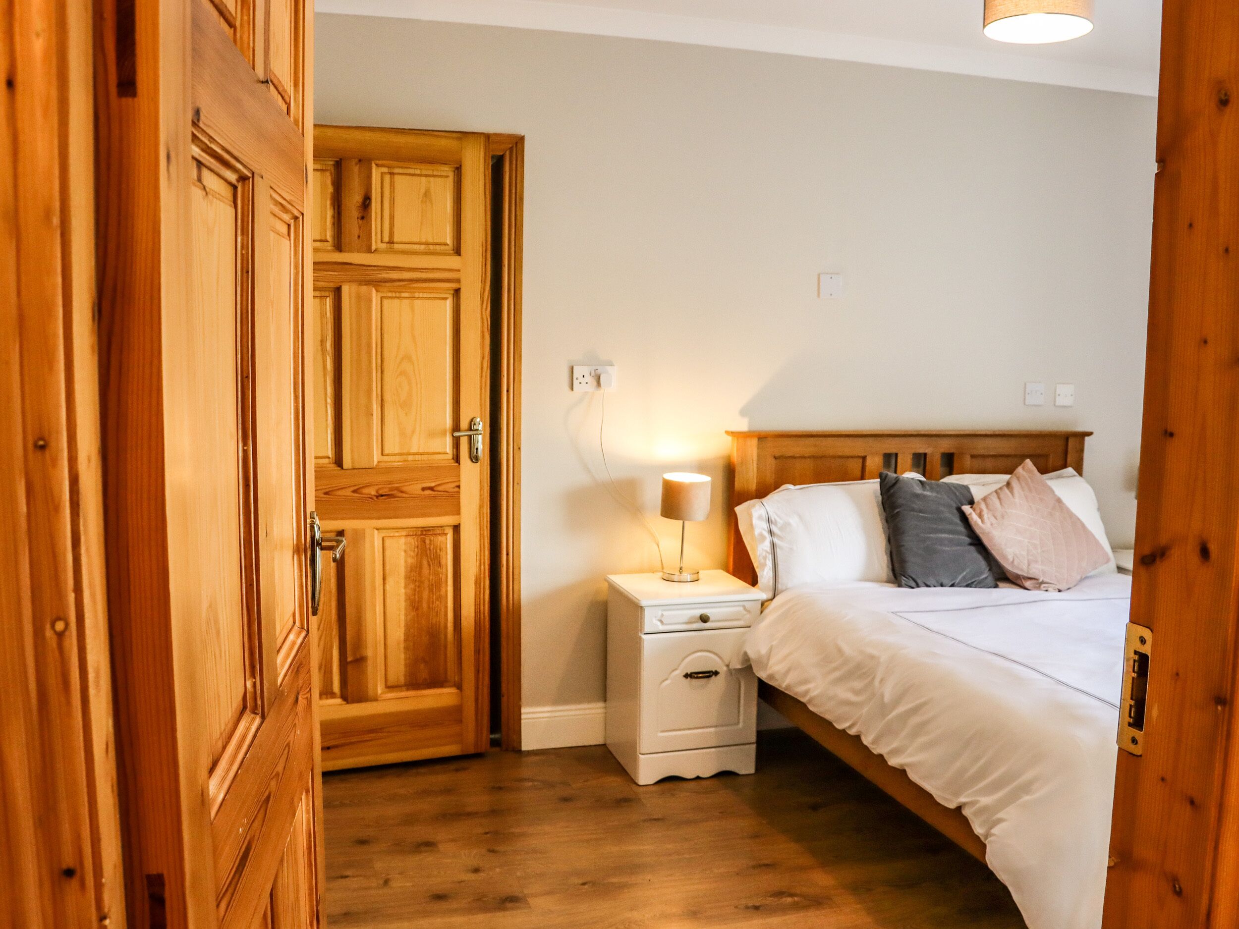 A bedroom with a wooden door, a bedside table with a lamp, a bed with pillows, and a nightstand.
