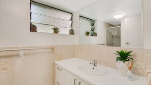 Une salle de bain avec un lavabo blanc, un miroir et une fenêtre située au-dessus du lavabo.
