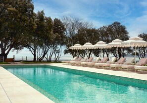 Zona de piscina con sillones reclinables y sombrillas, rodeada de árboles y con vistas a una masa de agua.