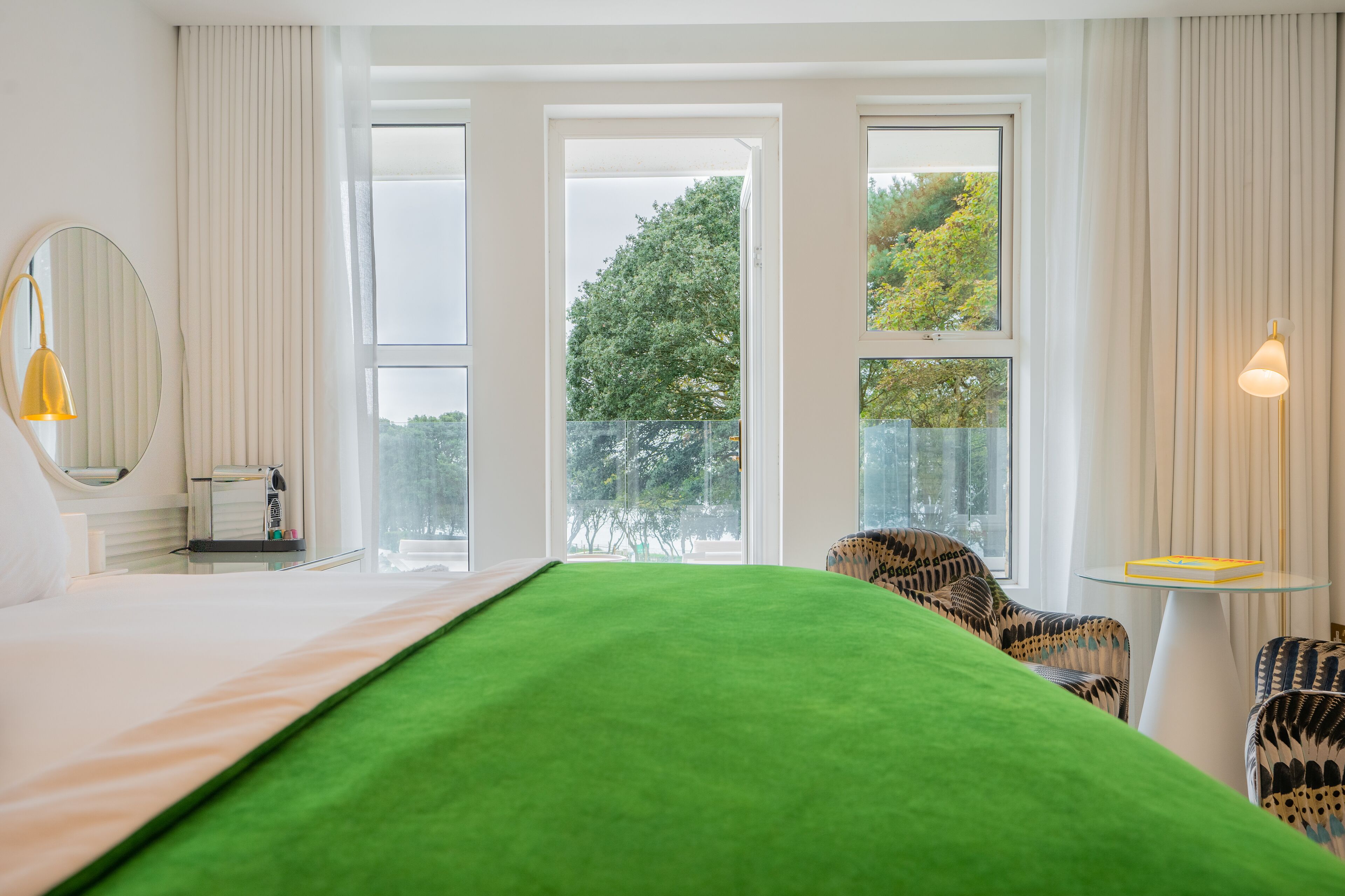 A hotel room with a large bed, a chair, a small table, and a window with a view of trees.