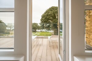 Een moderne kamer met een grote schuifpui die leidt naar een terras met houten vloer, een klein rond tafeltje en een bankje.