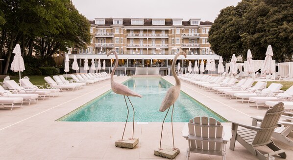 Zona de piscina con sillas de descanso y sombrillas, un edificio con balcones al fondo y dos esculturas de cisnes en la piscina.