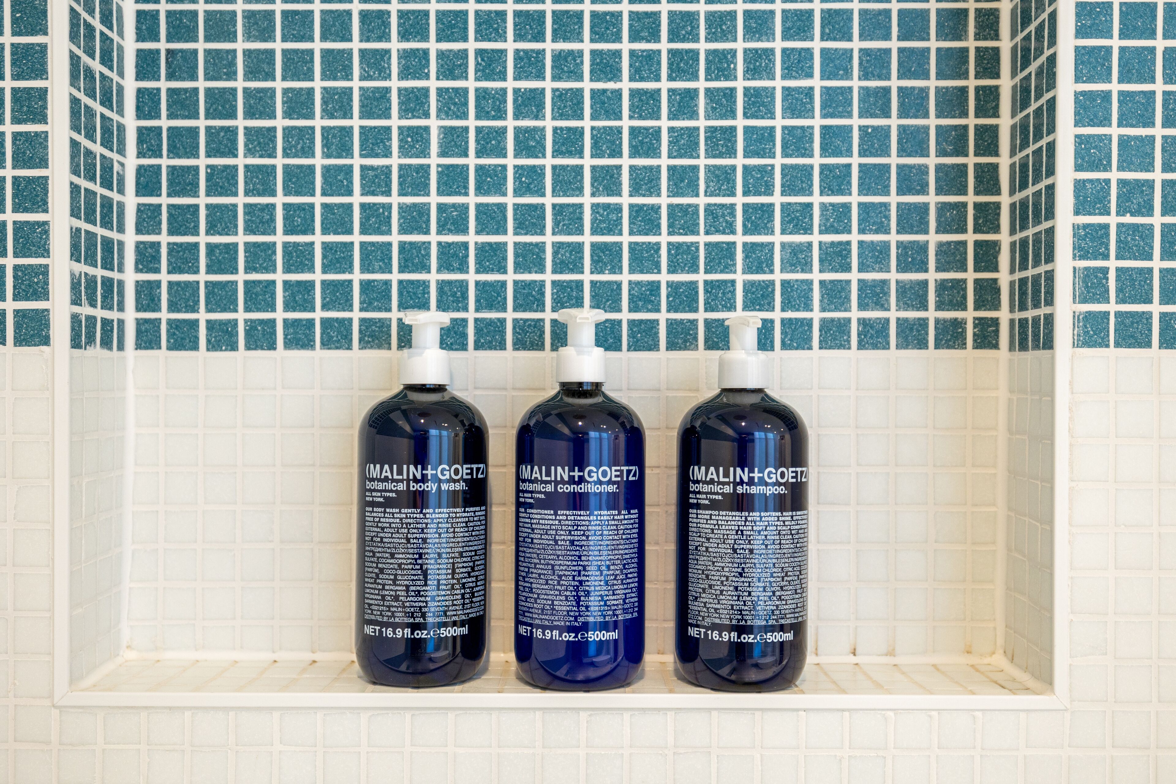 Tres botellas de gel de ducha corporal botánico MALIN+GOETZ sobre una estantería frente a una pared de azulejos.