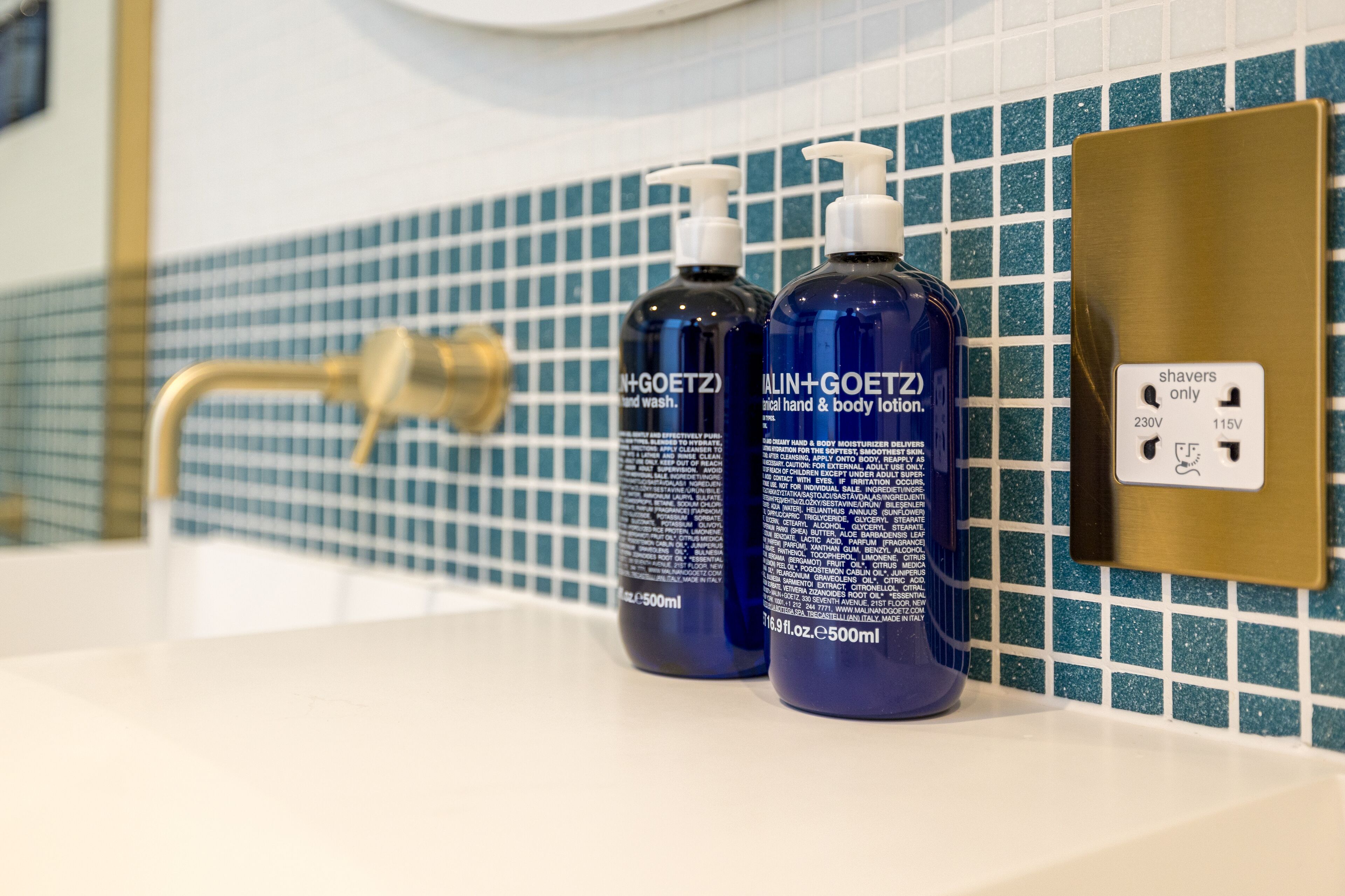 A bathroom with a white sink, two bottles of hand and body lotion, a gold faucet, and a blue tiled backsplash.