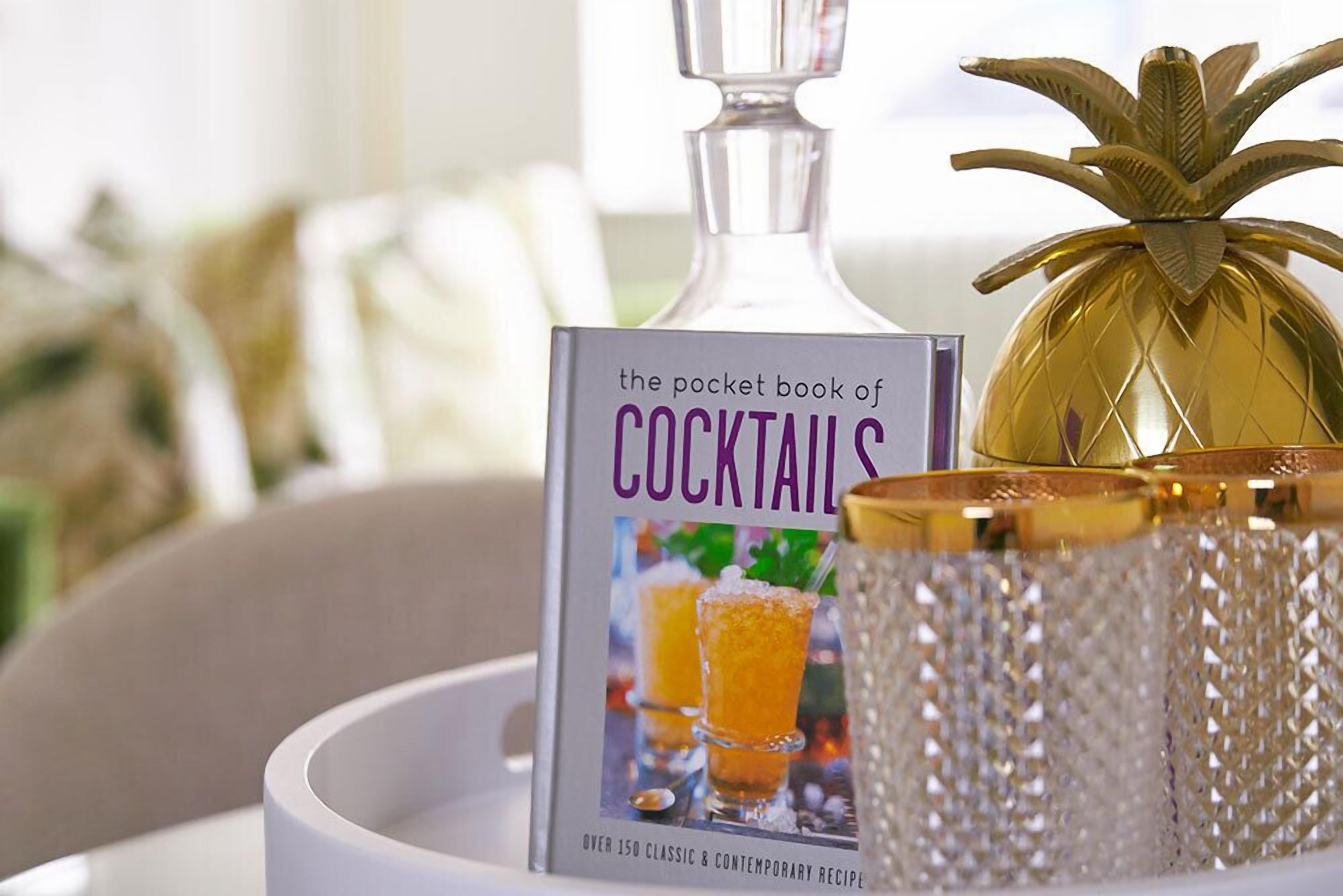 A book titled 'The Pocket Book of Cocktails' on a tray with a decorative pineapple and a glass container.