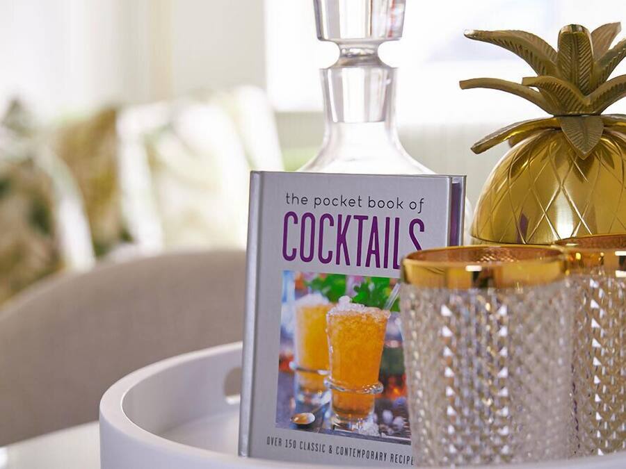 A book titled 'The Pocket Book of Cocktails' on a tray with a decorative pineapple and a glass container.