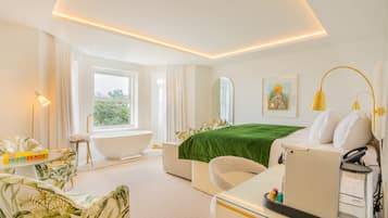 A modern hotel room with a large bathtub, a bed with a green bedspread, a sofa with a tropical print, and a desk with a lamp.