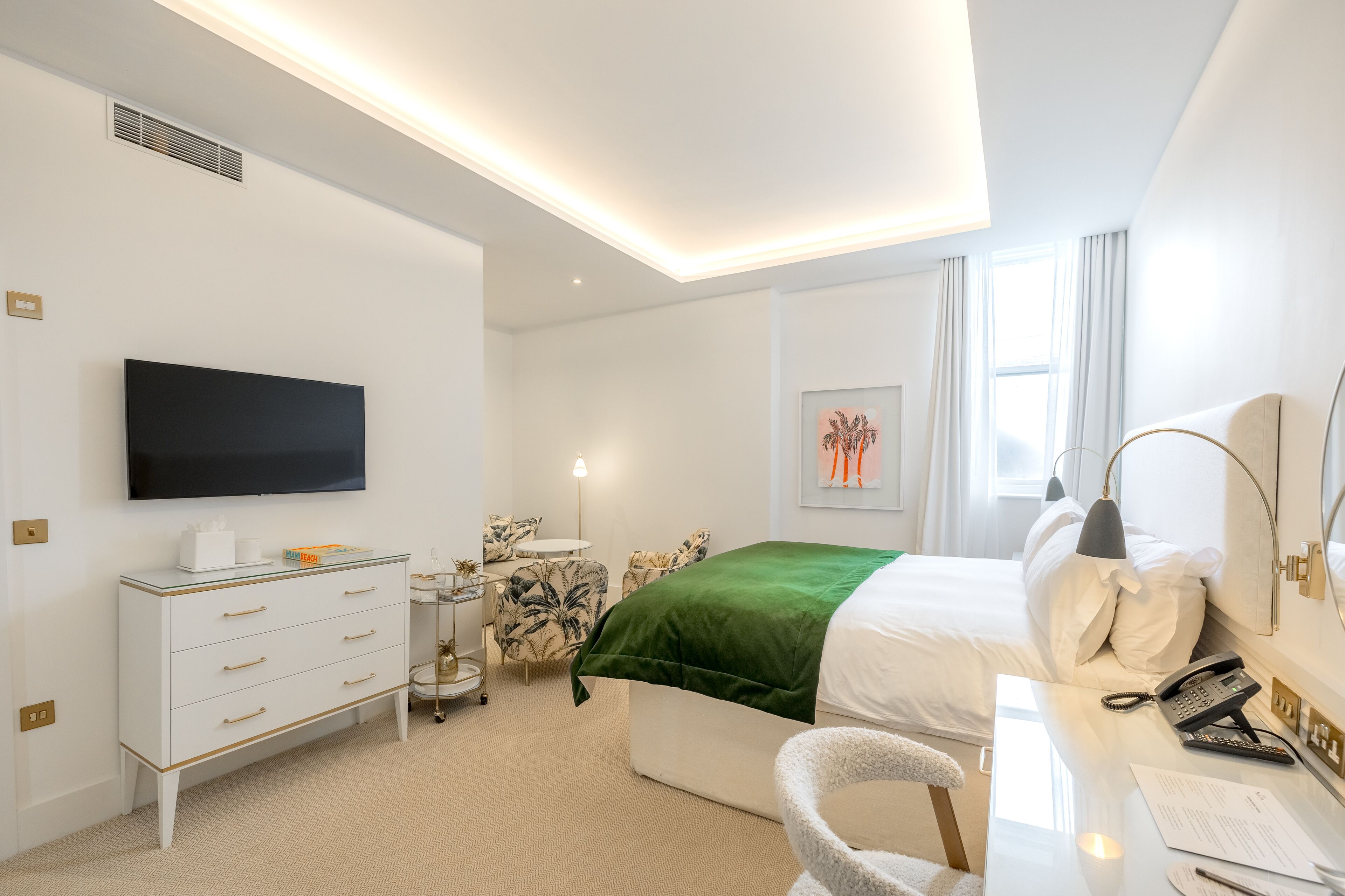 A modern hotel room with a flat-screen TV, a white dresser, a green bedspread, a desk with a phone, and a chair.