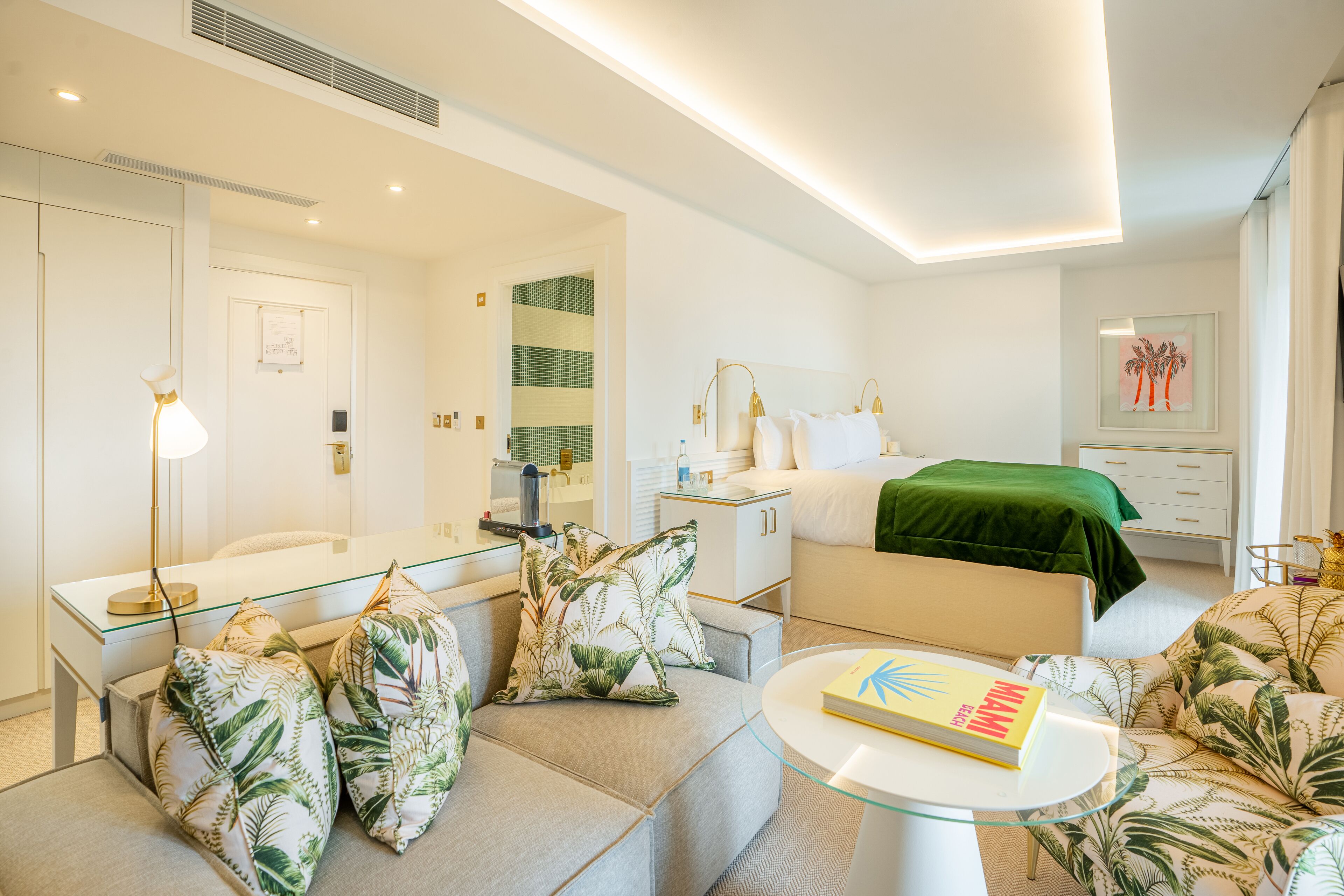 A modern hotel room with a glass coffee table, a sofa with tropical pillows, a bed with a green blanket, and a large mirror.