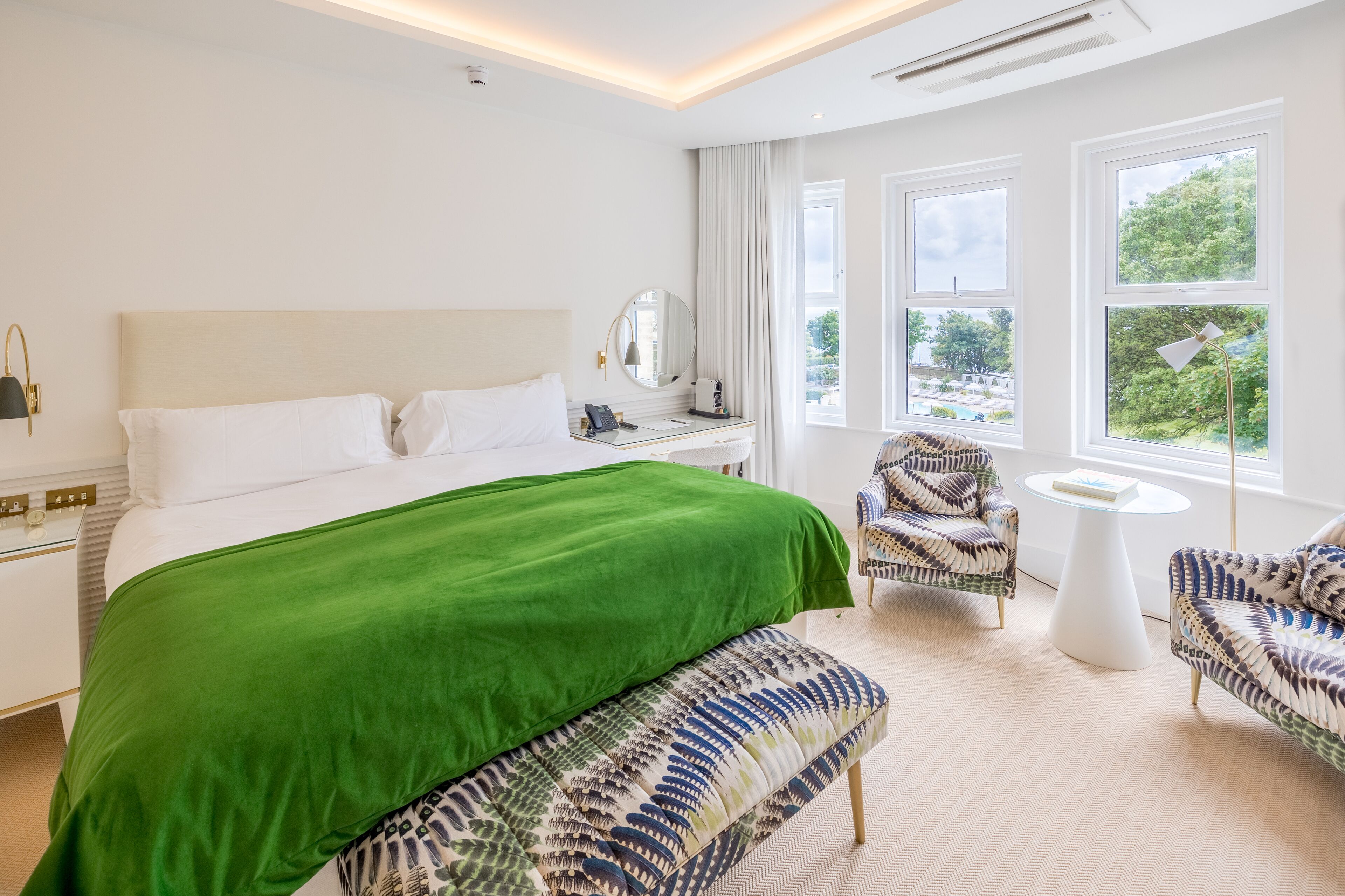 A hotel room with a large bed, a green throw, two patterned armchairs, a small round table, and a view of greenery through the windows.