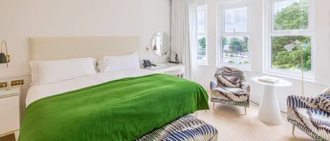 A hotel room with a large bed, a green throw, two patterned armchairs, a small round table, and a view of greenery through the windows.