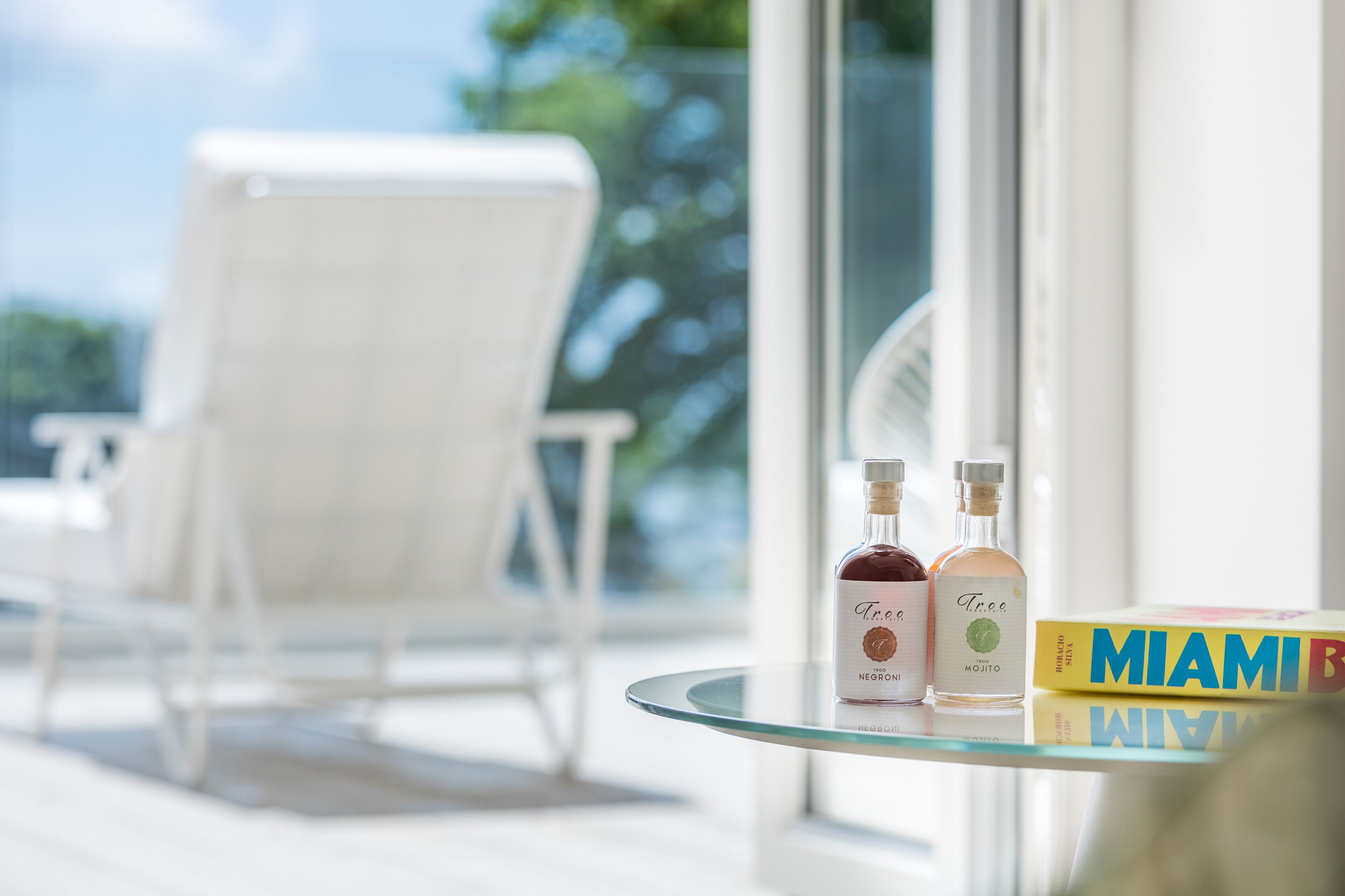 Ein Glastisch mit zwei alkoholischen Getränken und einem Buch mit dem Titel "MIAMI" auf einem Balkon mit einem weißen Stuhl.