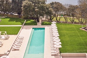 Una piscina rectangular con sillones blancos para tomar el sol y un área de césped con árboles.