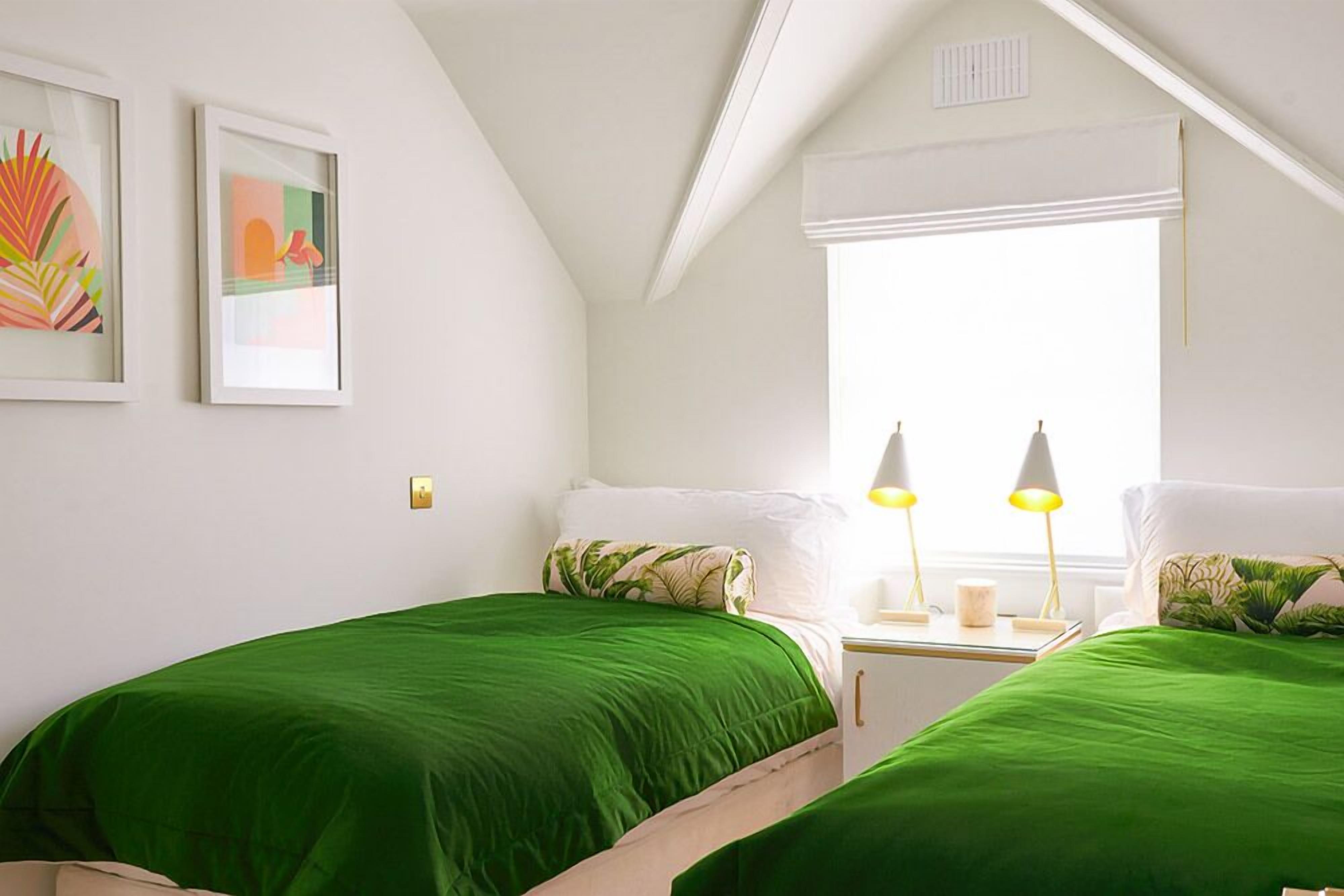 Un dormitorio con dos camas, ropa de cama verde, una ventana con contraventanas blancas y dos lámparas de noche.