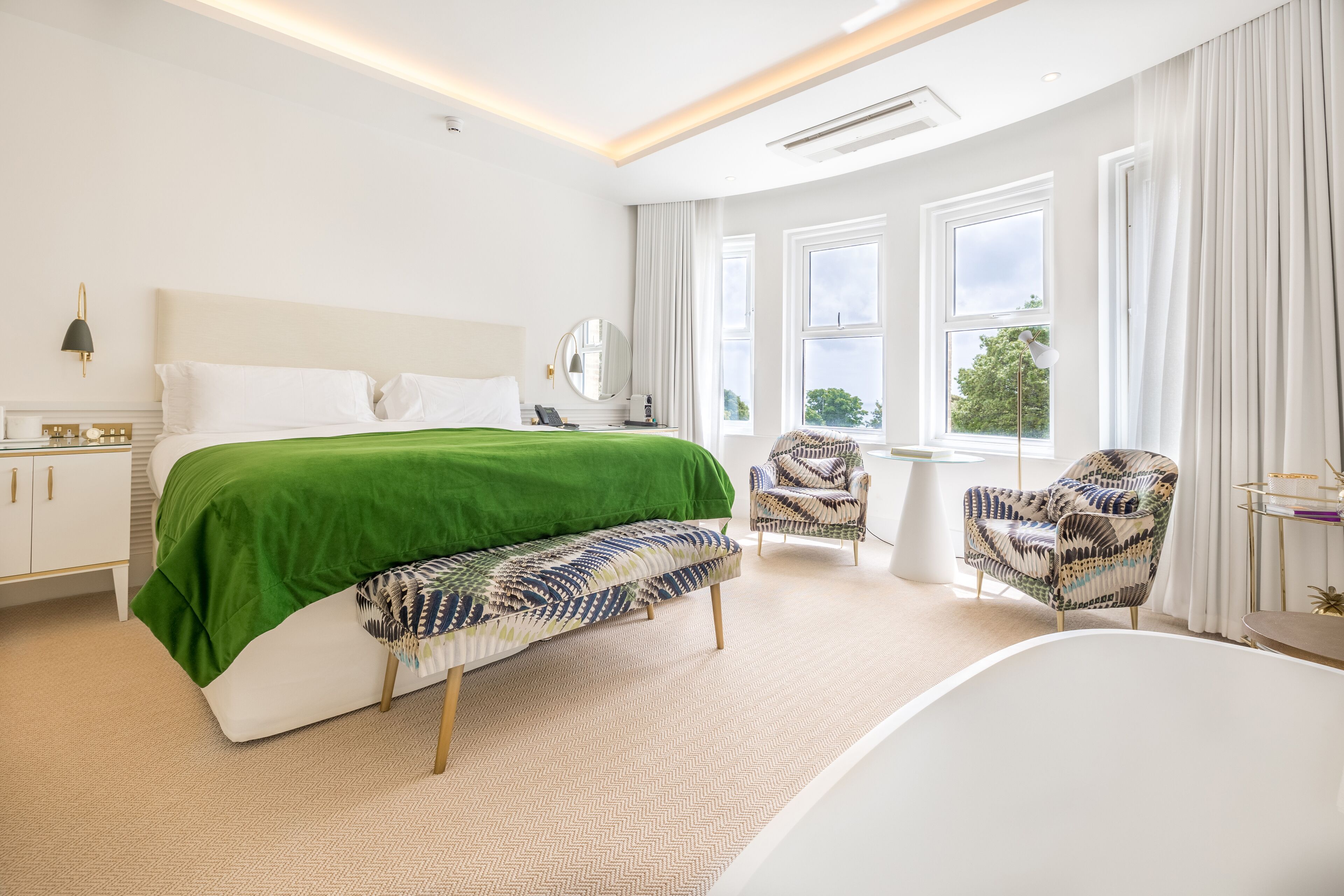 A modern hotel room with a large bed, a bench, a chair, a desk, and a bathtub.