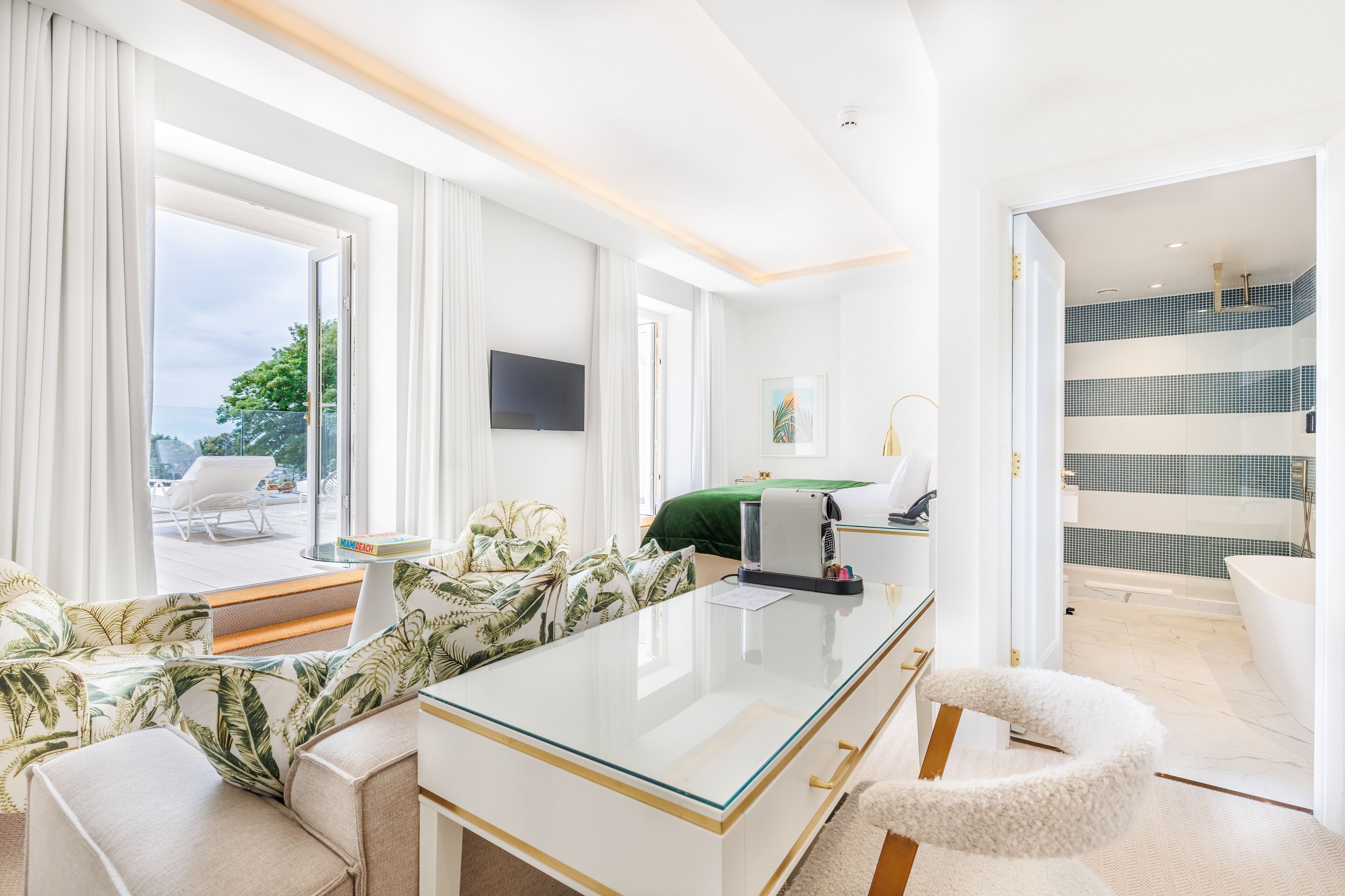A modern hotel room with a glass desk, a sofa with a tropical print, a flat-screen TV, and a view of the outdoors.