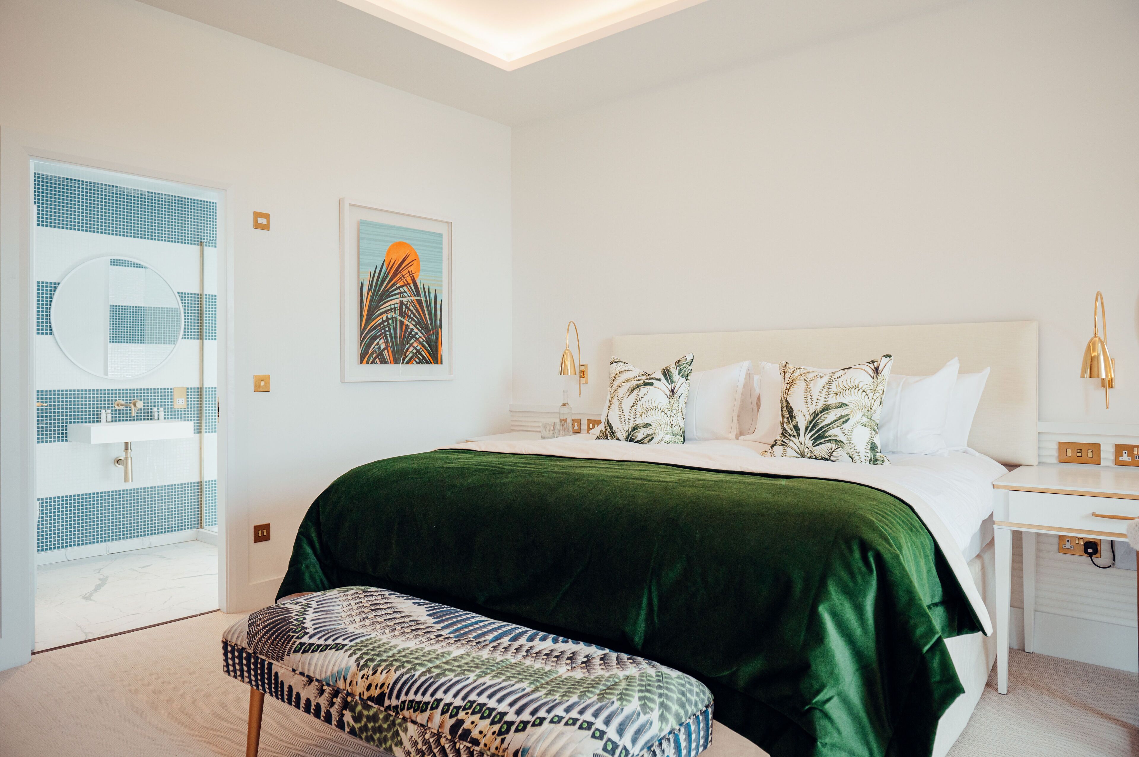 A modern bedroom with a large bed, a green velvet bedspread, a patterned bench, a white bedside table with a lamp, and a wall art piece.