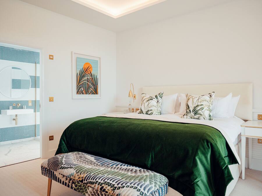 A modern bedroom with a large bed, a green velvet bedspread, a patterned bench, a white bedside table with a lamp, and a wall art piece.