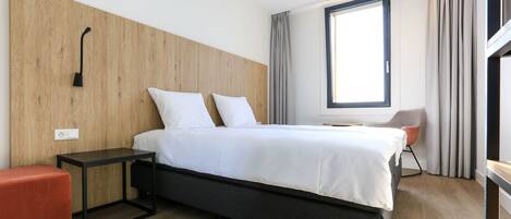 Habitación de hotel moderna con cabecera de madera, cama con ropa blanca, una mesita auxiliar pequeña, un reposapiés rojo, una ventana con cortinas y una silla.