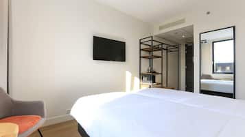 A modern hotel room with a large bed, a flat-screen TV mounted on the wall, a wooden chair, and a large mirror reflecting the room.