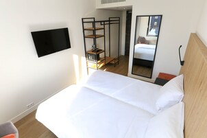 Una habitación de hotel con una cama, un televisor montado en la pared, un mueble estante de madera y un espejo que refleja la habitación.