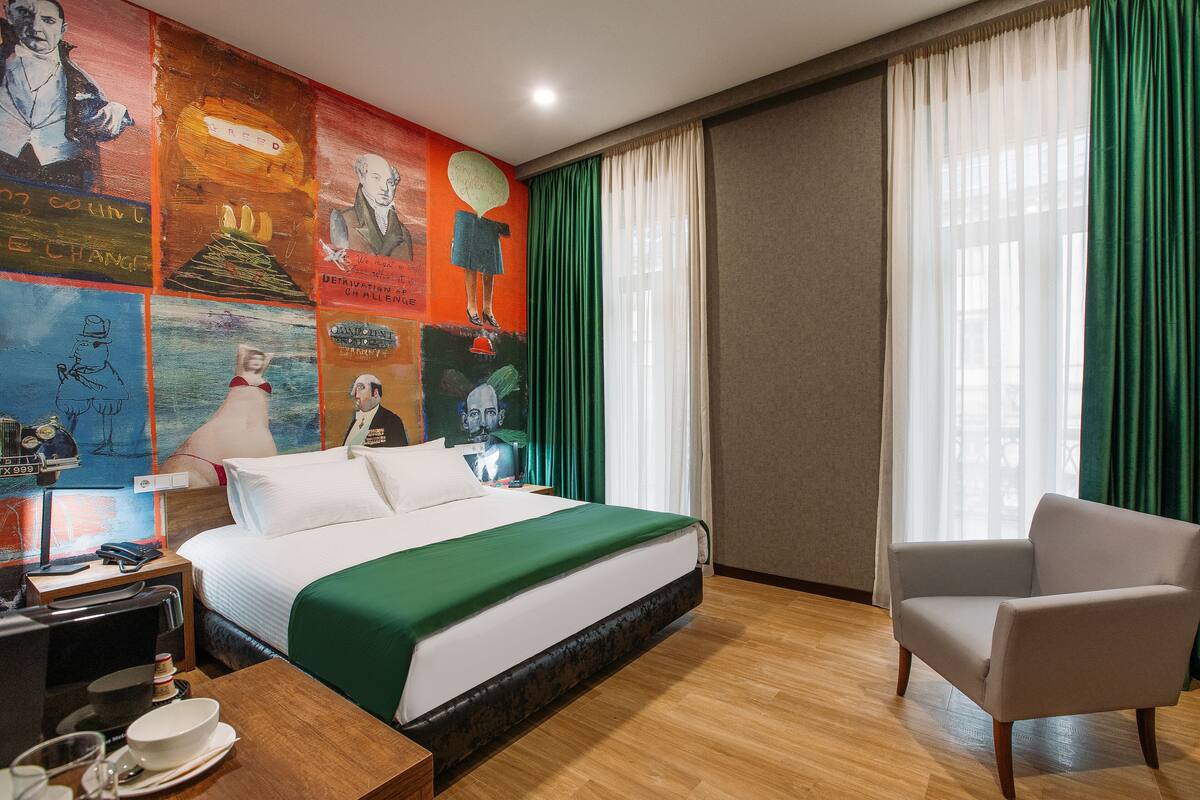 A hotel room with a bed, a chair, a desk with a phone and a lamp, and a wall with colorful artwork.