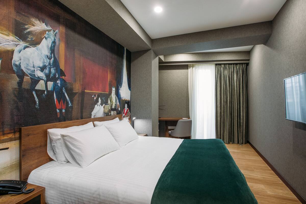 A hotel room with a large bed, a desk, and a chair. There is a painting on the wall and a TV on the wall.