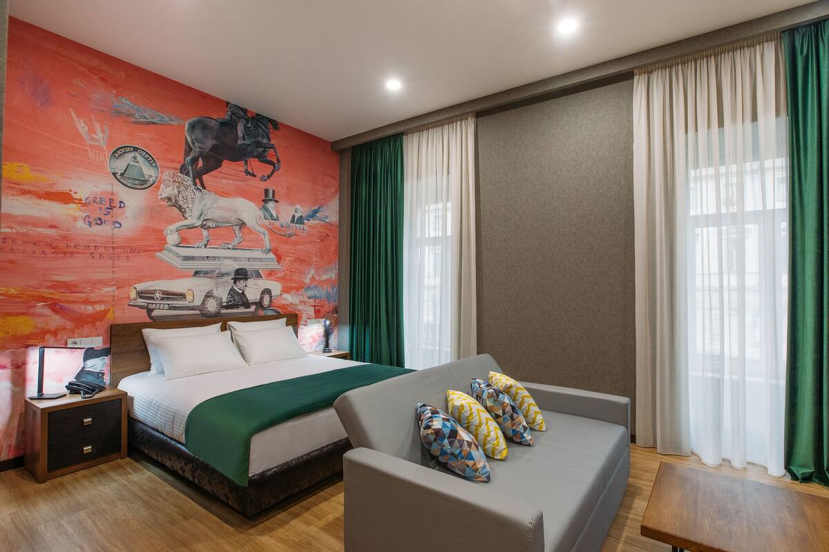 A hotel room with a bed, a sofa, a desk, and a mural on the wall.