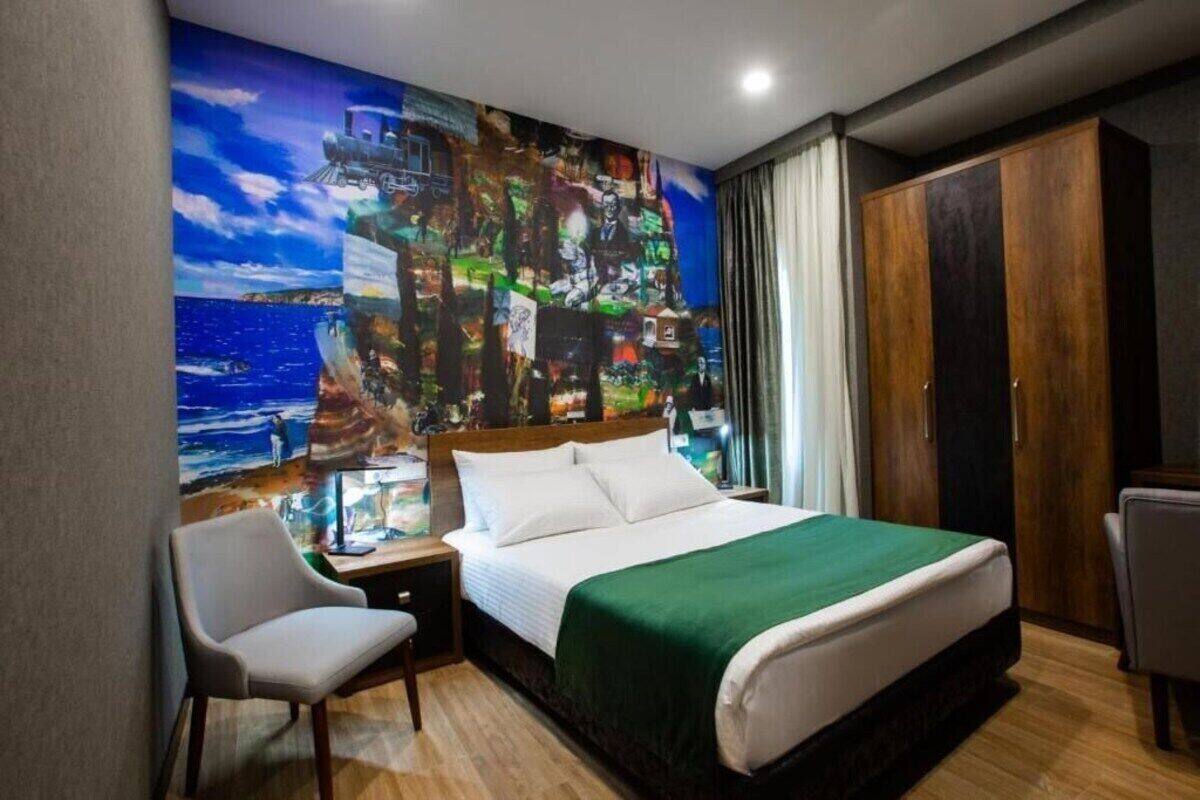 A hotel room with a large bed, a desk with a chair, a wardrobe, and a colorful abstract mural on the wall.