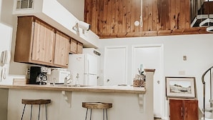 A kitchen with wooden cabinets, a white countertop, two stools, a coffee maker, and a framed picture on the wall.
