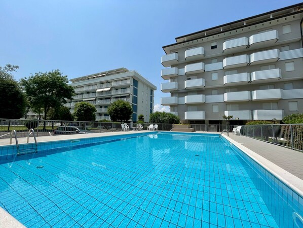 A swimming pool with tiled flooring, surrounded by a metal fence. In the background, there is a multi-story building with balconies.
