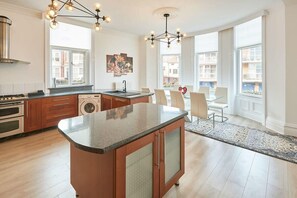 A modern kitchen with granite countertops, a built-in oven, and a large island. There is a dining area with a glass table and chairs, and a large window providing a view of the city. - Host & Stay | Sea Forever Seafront Villa (Saltburn-by-the-Sea)