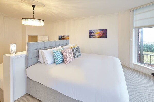 A spacious bedroom with a large bed, a bedside table, a chandelier, and a window with a view. - Host & Stay | Sea Forever Seafront Villa (Saltburn-by-the-Sea)