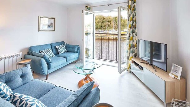 Una sala de estar moderna con un sofá azul, una mesa de centro de vidrio, un televisor sobre un mueble de madera y un balcón con vista a la calle.