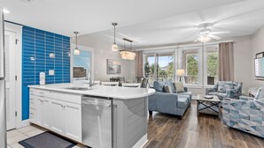 A modern kitchen with a central island, a dining area with a blue sofa, and a living room with a patterned sofa. - Rates Reduced! Oversized Balcony, Lagoon Pool Views! Hot Tub, Beach Tram, Gym (Miramar Beach)