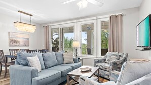 A living room with a sofa, armchairs, a coffee table, and a TV. There are large windows with curtains, a ceiling fan, and a chandelier. - Rates Reduced! Oversized Balcony, Lagoon Pool Views! Hot Tub, Beach Tram, Gym (Miramar Beach)