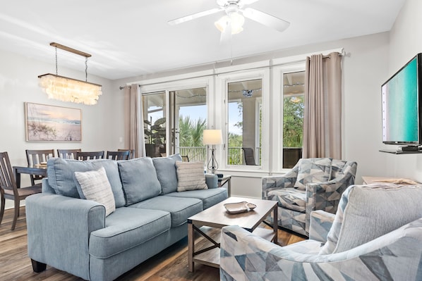Un salon comprenant un canapé, des fauteuils, une table basse et un téléviseur. De grandes fenêtres sont présentes, ainsi que des rideaux, un ventilateur de plafond et un lustre.
