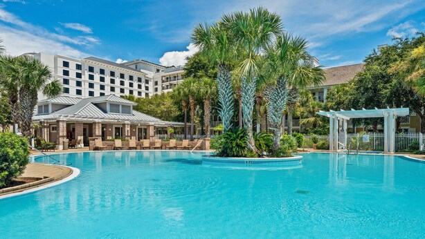 A large outdoor swimming pool surrounded by palm trees and a hotel building with multiple floors and balconies. - Rates Reduced! Oversized Balcony, Lagoon Pool Views! Hot Tub, Beach Tram, Gym (Miramar Beach)