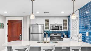 A modern kitchen with white cabinets, a stainless steel refrigerator, a microwave, and a stove. There are two white bar stools and pendant lights above the counter. - Rates Reduced! Oversized Balcony, Lagoon Pool Views! Hot Tub, Beach Tram, Gym (Miramar Beach)