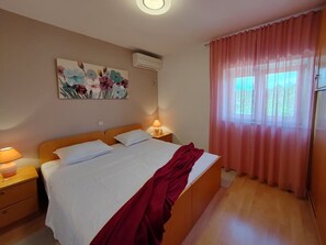 A bedroom with a bed, a nightstand, an air conditioner, a window with curtains, and a floral painting on the wall.