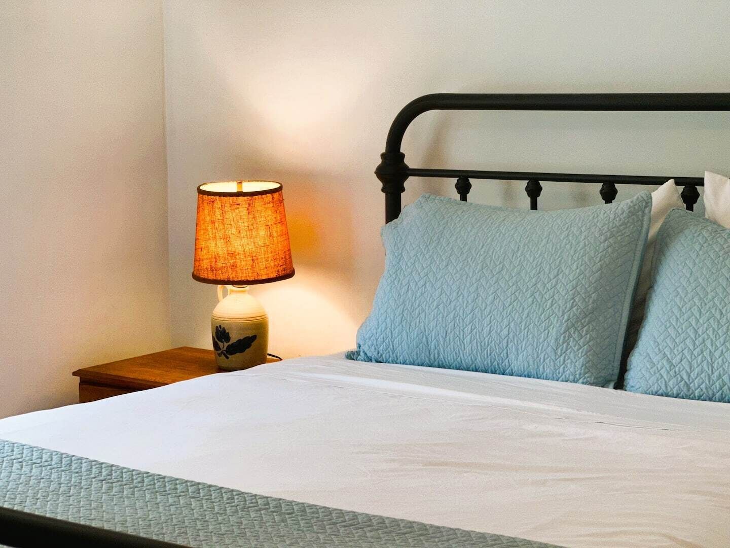 Un lit bien fait, avec un drap blanc et deux oreillers bleu clair, une table de chevet avec une lampe et une tête de lit en métal noir.