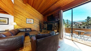 L’intérieur chaleureux d’un chalet en bois, avec une cheminée en pierre, un canapé en cuir et un balcon offrant une vue imprenable sur les montagnes.