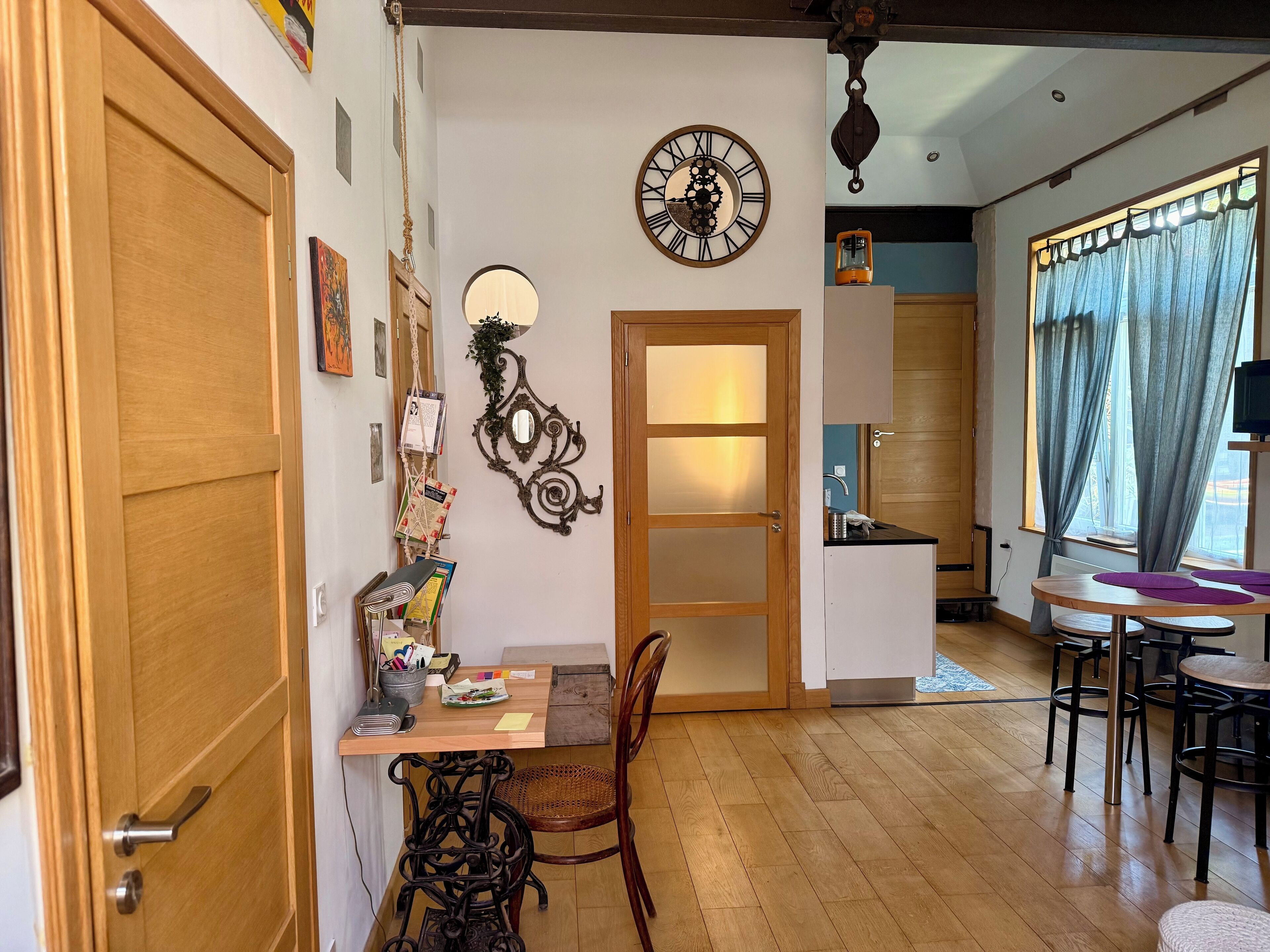 Een gezellige kamer met een houten deur, een wandklok, een zithoek met een tafel en stoelen, en een raam met gordijnen.