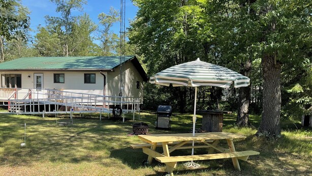 Een picknicktafel met een gestreepte parasol, een barbecue en een kleine schuur in een gazon, met een huis op de achtergrond.